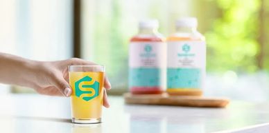 Hand holding a glass of juice with bottles blurred in the background.