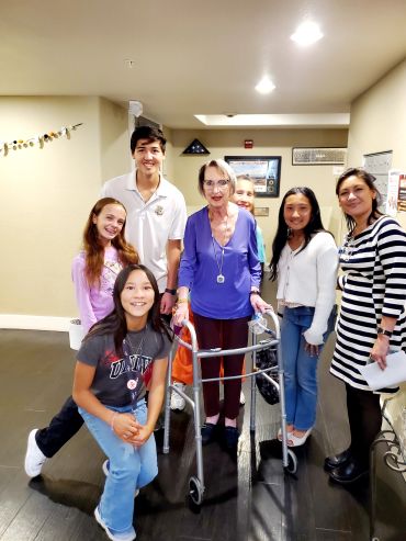 A diverse group of people posing together indoors, including an elderly woman with a walker.