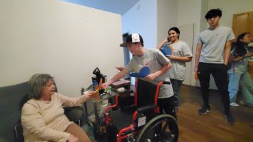 A boy in a wheelchair handing a card to an elderly woman on a couch.