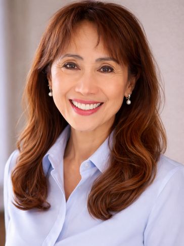 Smiling woman with long brown hair wearing a light blue shirt and pearl earrings.