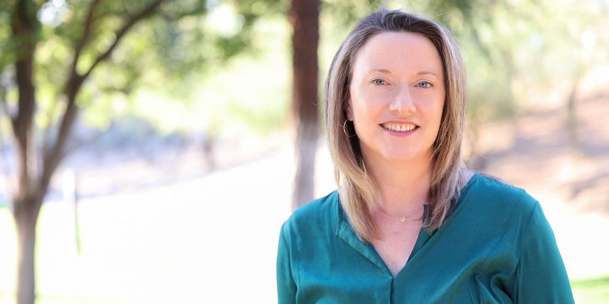 Smiling woman in a teal blouse outdoors with trees in the background.