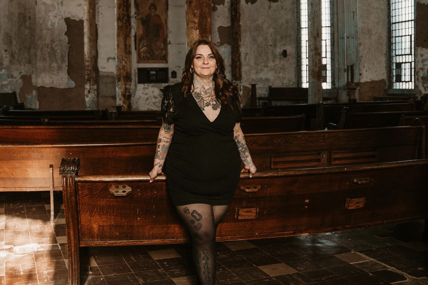 Woman with tattoos in a black dress leaning on a church pew.