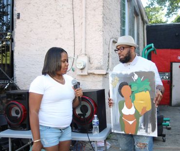 A man shows a painting to a woman holding a microphone outdoors.
