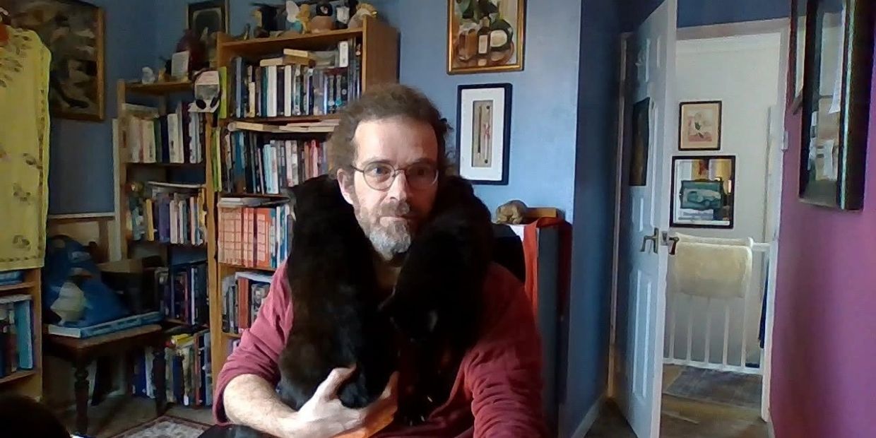 Man with glasses holds two black cats around his neck.
