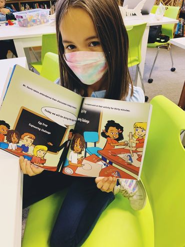 Young girl wearing a mask reads a colorful book in a classroom.