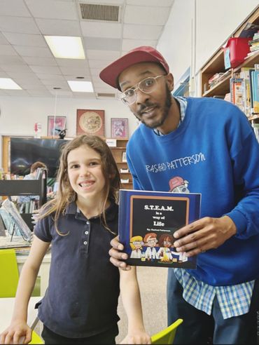 A man and a girl smiling in a library holding a book titled 'S.T.E.A.M. is a way of Life'.
