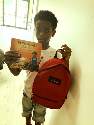 Boy holding a book titled 'Slow Money is Better Than No Money' and a red backpack.