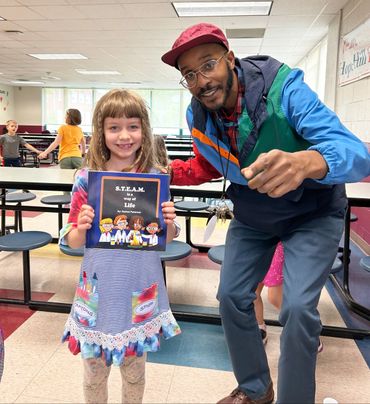 A young girl proudly holds a S.T.E.A.M. book with an adult beside her in a school cafeteria.
