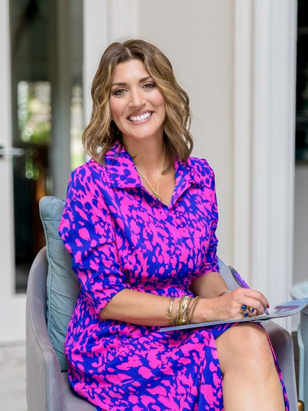 Smiling woman in a vibrant pink and blue dress sitting and writing in a notebook.