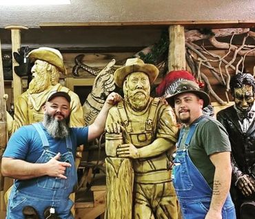 Two men in overalls posing with wooden statues of lumberjacks.