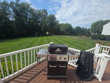 A grill on a wooden deck overlooking a large, striped lawn with trees and a playset.
