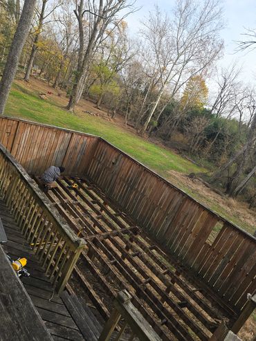 Person working on a wooden deck frame in a backyard surrounded by trees.