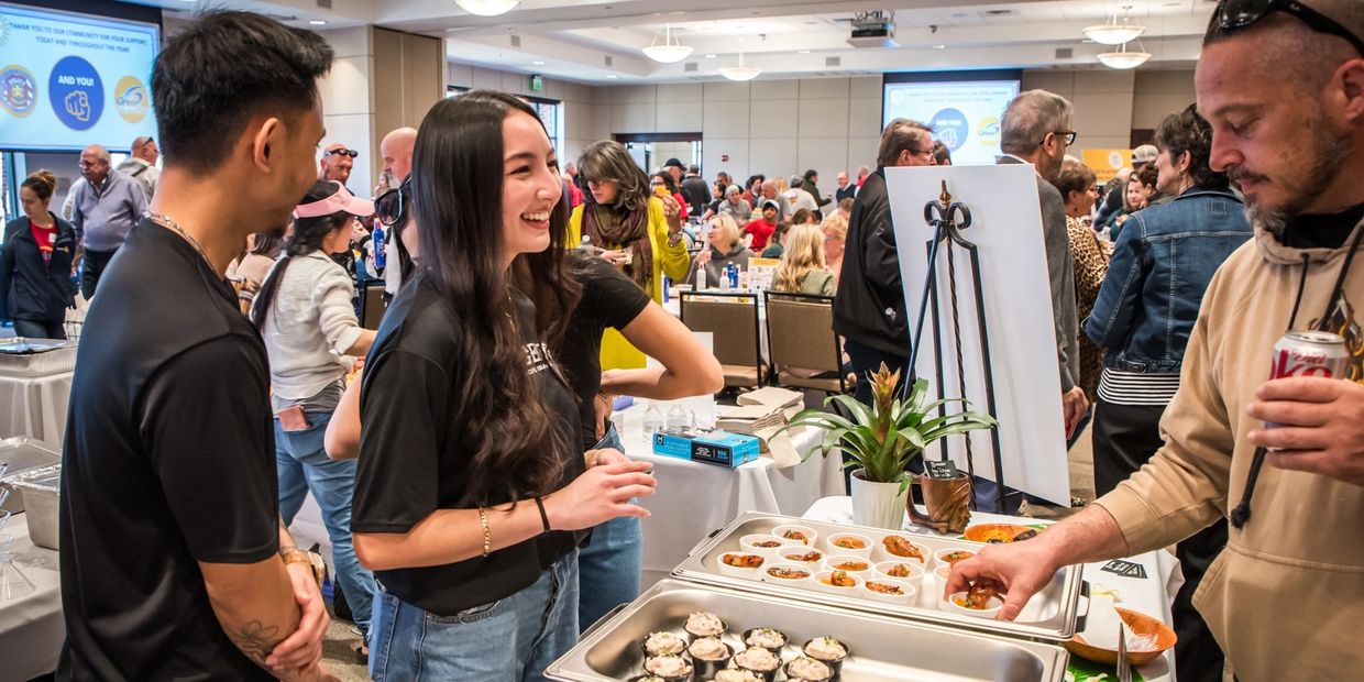 Young women smiling and serving food, background is room full of people eating.
