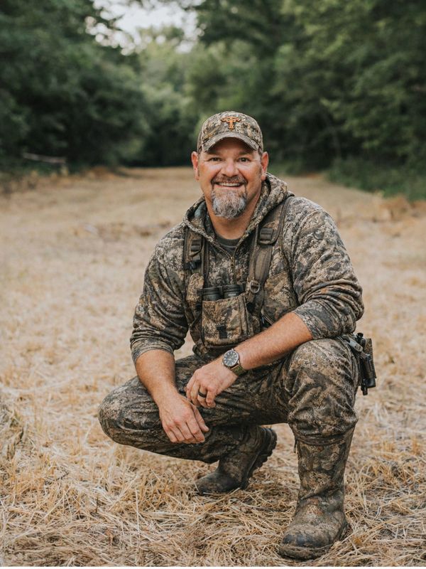 Man in camouflage hunting gear smiling while crouching outdoors.