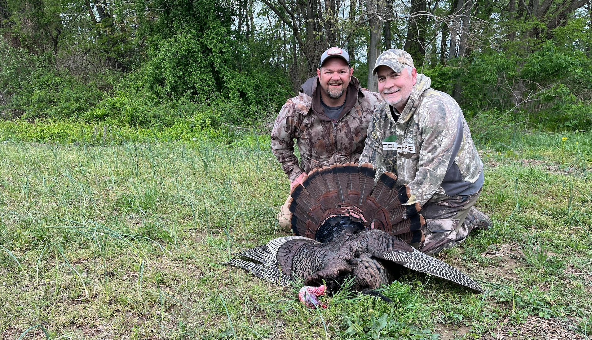Two hunters proudly display a wild turkey they caught in a forest clearing.