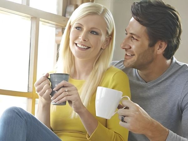 Couple by window, holding coffee mugs.
