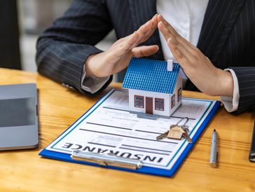 Hands protecting a house model over insurance papers.