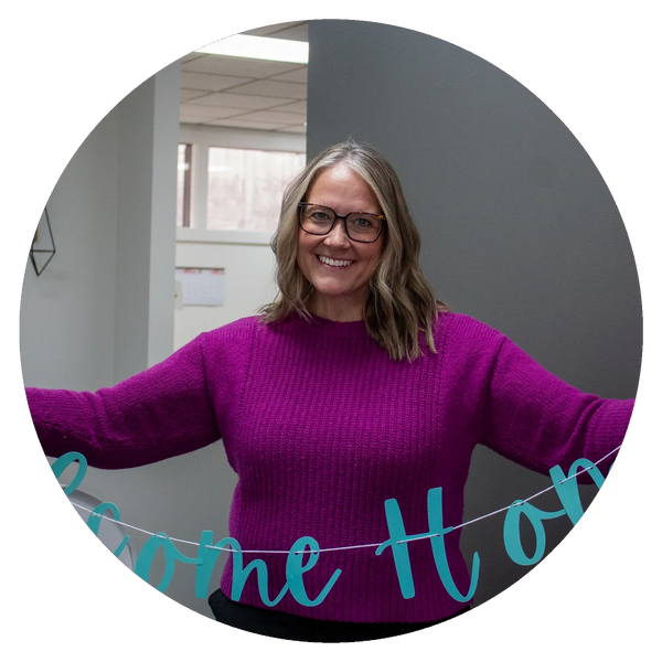 Woman in purple sweater smiling and holding a "Welcome Home" sign indoors.