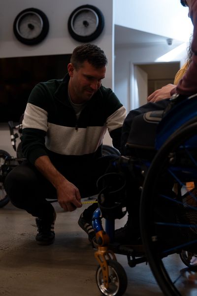Man kneeling and adjusting a wheelchair for another person indoors.