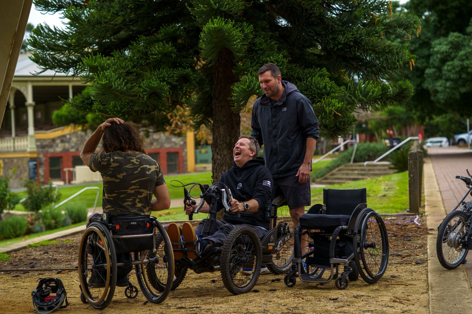 Three men, two in wheelchairs, enjoying a lively conversation outdoors under a large tree.