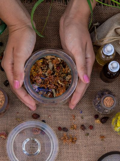 A pair of hands holding a jar of dried herbs and flowers with other herbal items on a burlap surface