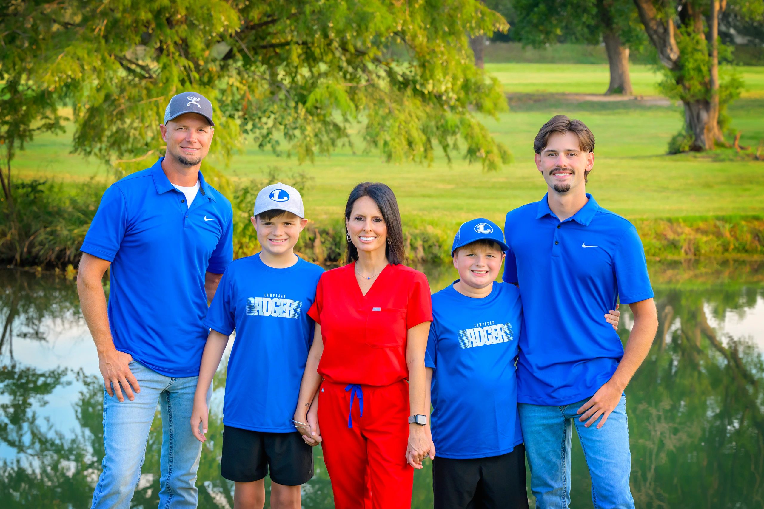 A family stands together outdoors near a pond, smiling and dressed casually.