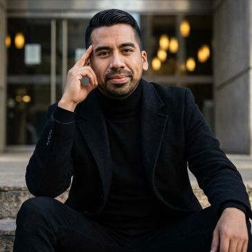 Confident man in black outfit sitting outdoors with thoughtful expression.