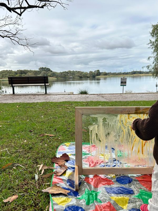 Person painting on a transparent canvas by a lakeside on a colorful mat.