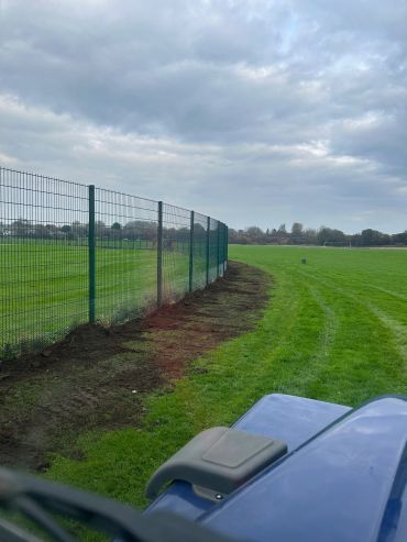 Lawn mowing next to a fenced sports field under a cloudy sky.