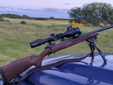Rifle with scope resting on a vehicle in an open field.
