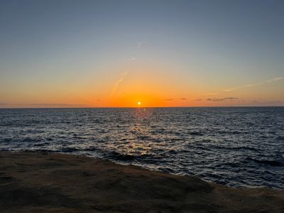 Sun setting over calm ocean waters with rocky shore foreground.