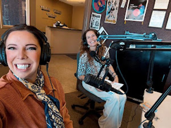 Two women smiling in a radio broadcasting studio with microphones and headphones.