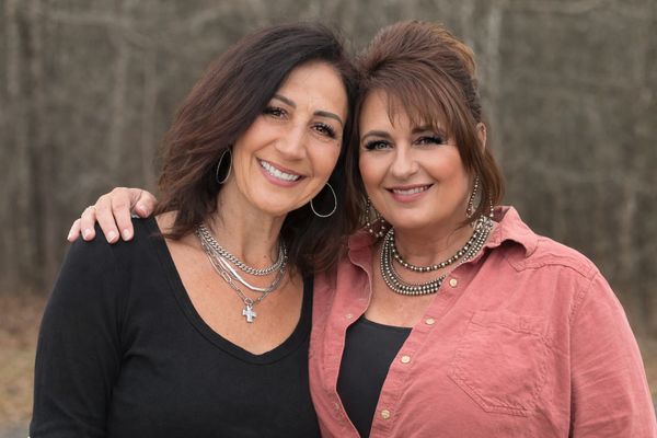 Two women smiling warmly, embracing outdoors with trees in the background.