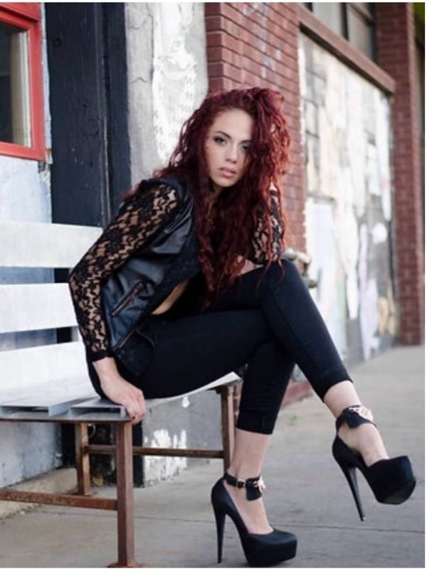 Woman with curly red hair sitting on a bench wearing black lace and leather outfit with high heels in a high fashion look.
