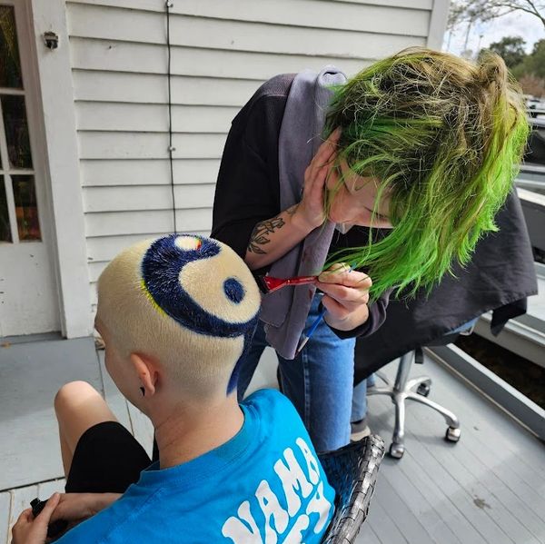 stylist with green hair painting a yin-yang design onto the shaved head of a client outdoors