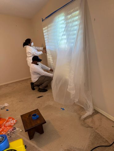 Two people in protective gear taping plastic sheeting to a wall indoors.
