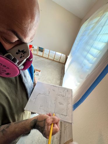 Person wearing a mask holding a notebook with a room layout sketch indoors.