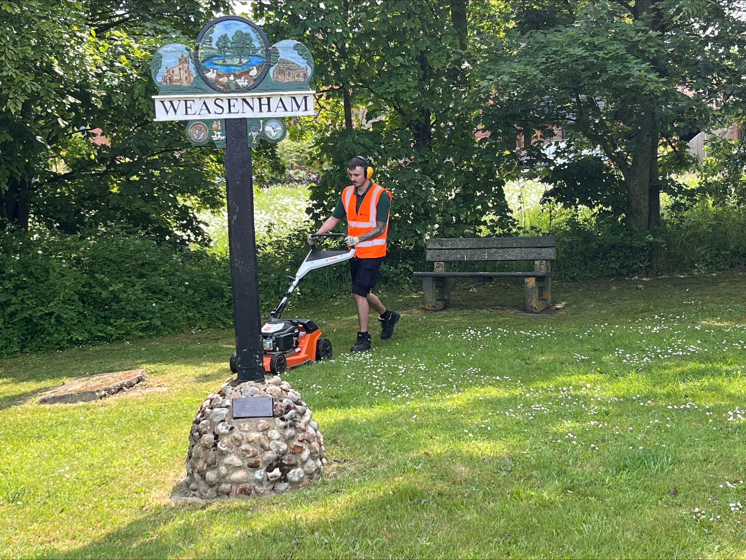 Gardener in a hi vis jacket with a petrol lawn mower cutting the grass around the Weasenham sign.