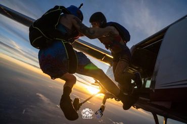 Two skydivers jumping from a plane at sunset, holding hands mid-air.