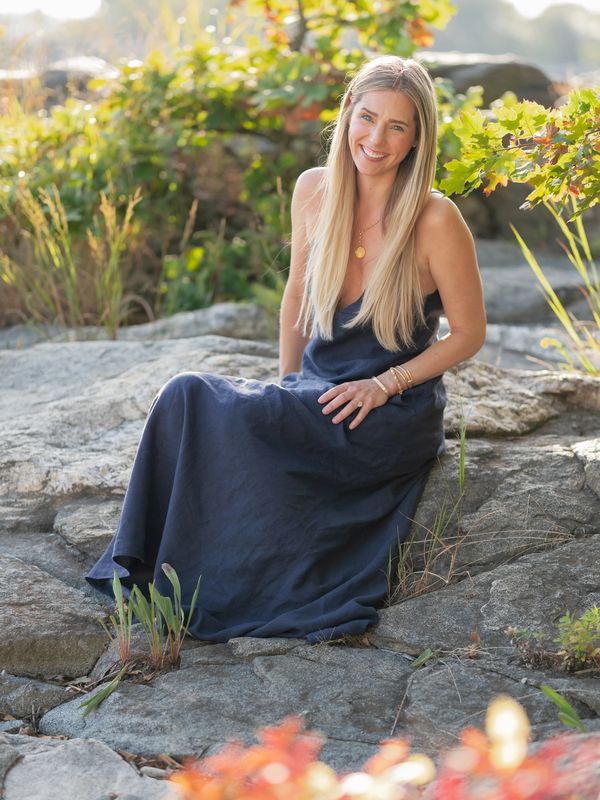 Smiling woman in a navy dress sitting on rocks in a sunny garden.
