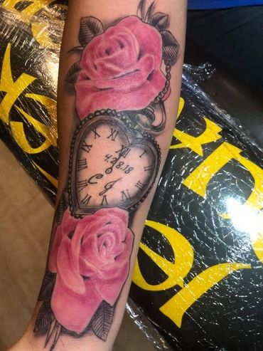 Heart-shaped clock tattoo with pink roses and initials on forearm.