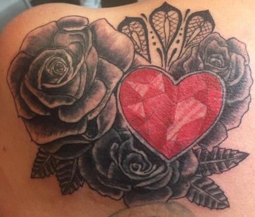 Tattoo of a red faceted heart surrounded by black and gray roses on skin.
