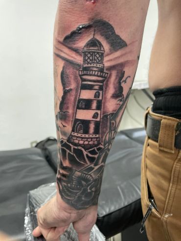 Black and gray lighthouse tattoo on forearm with clouds and birds.