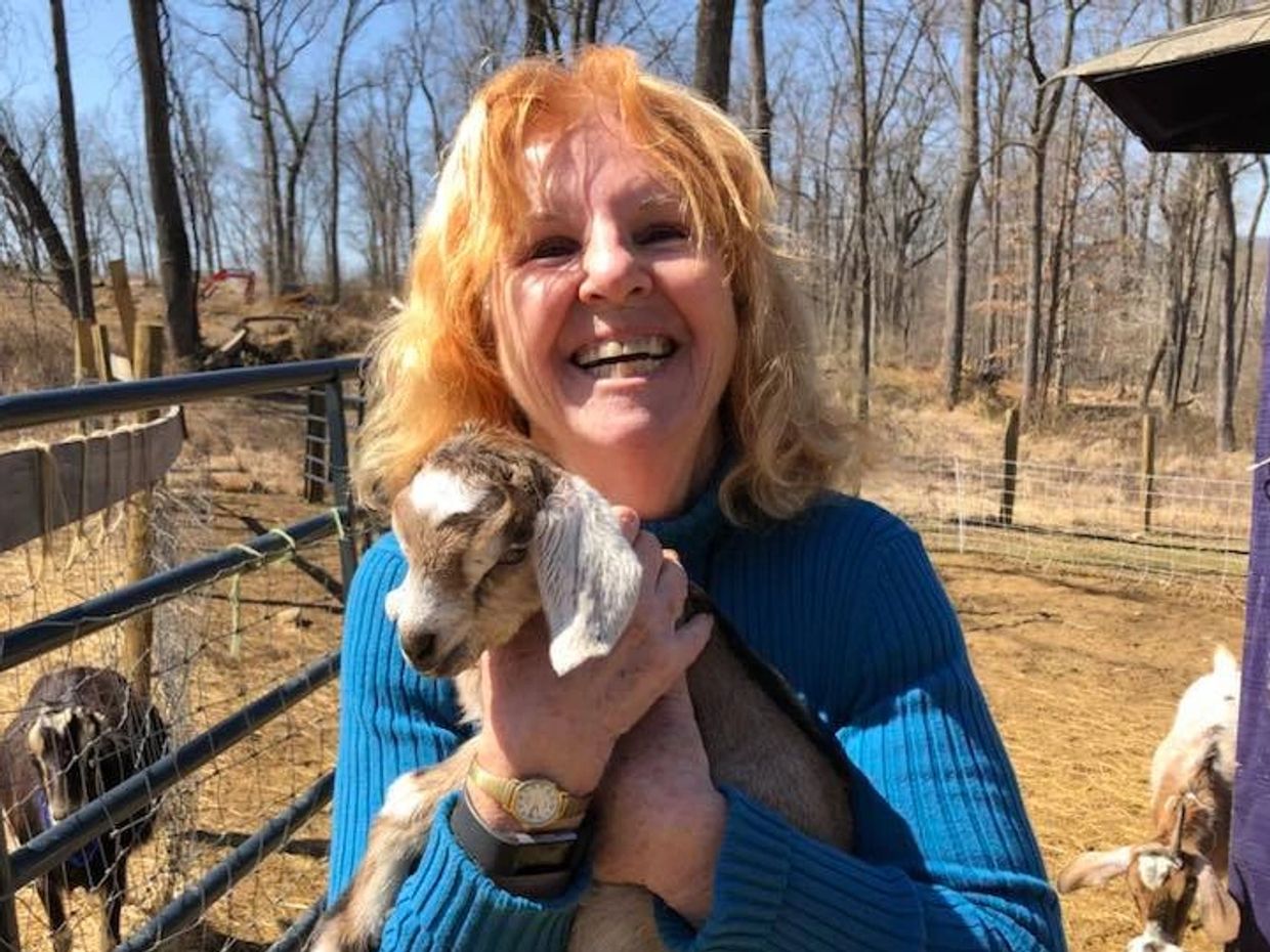 A smiling, red-headed Kathleen wears a blue sweater and hugs a baby goat kid.