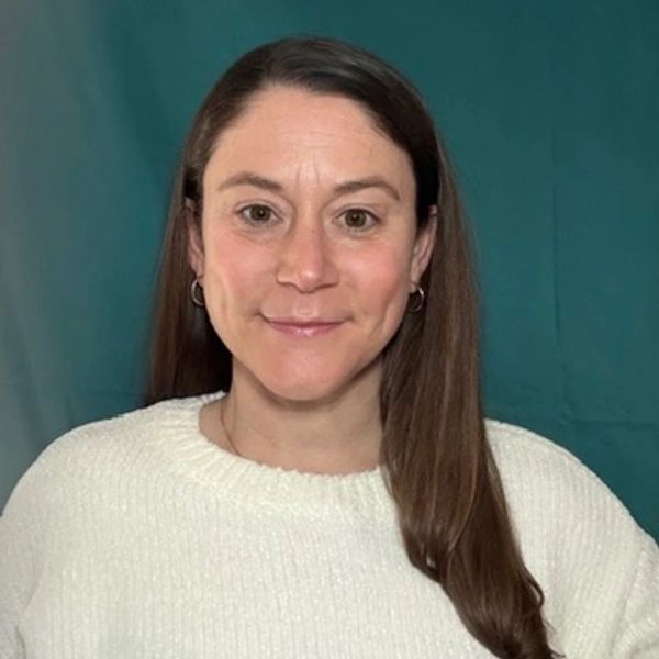 Smiling woman with long brown hair wearing a white sweater against a teal background.