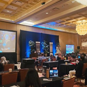 Speaker presenting at a conference in a hotel ballroom with attentive audience.