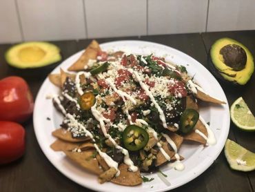 Plate of loaded nachos with jalapeños, crema, and cheese, surrounded by fresh avocado, tomatoes, and lime.