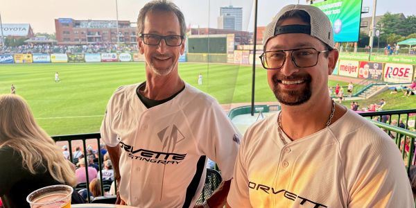 Two men smiling at a baseball game, wearing matching Corvette shirts.