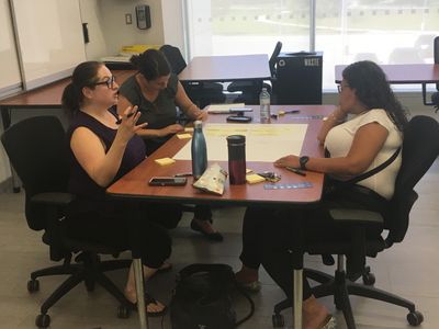 Three women sit talking at a table with paper and water bottles on it.