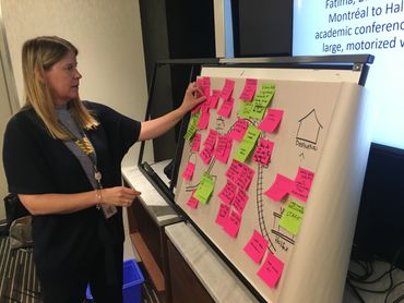 A woman stands in front of a bunch of pink and green post-it notes stuck to a large pad of paper.